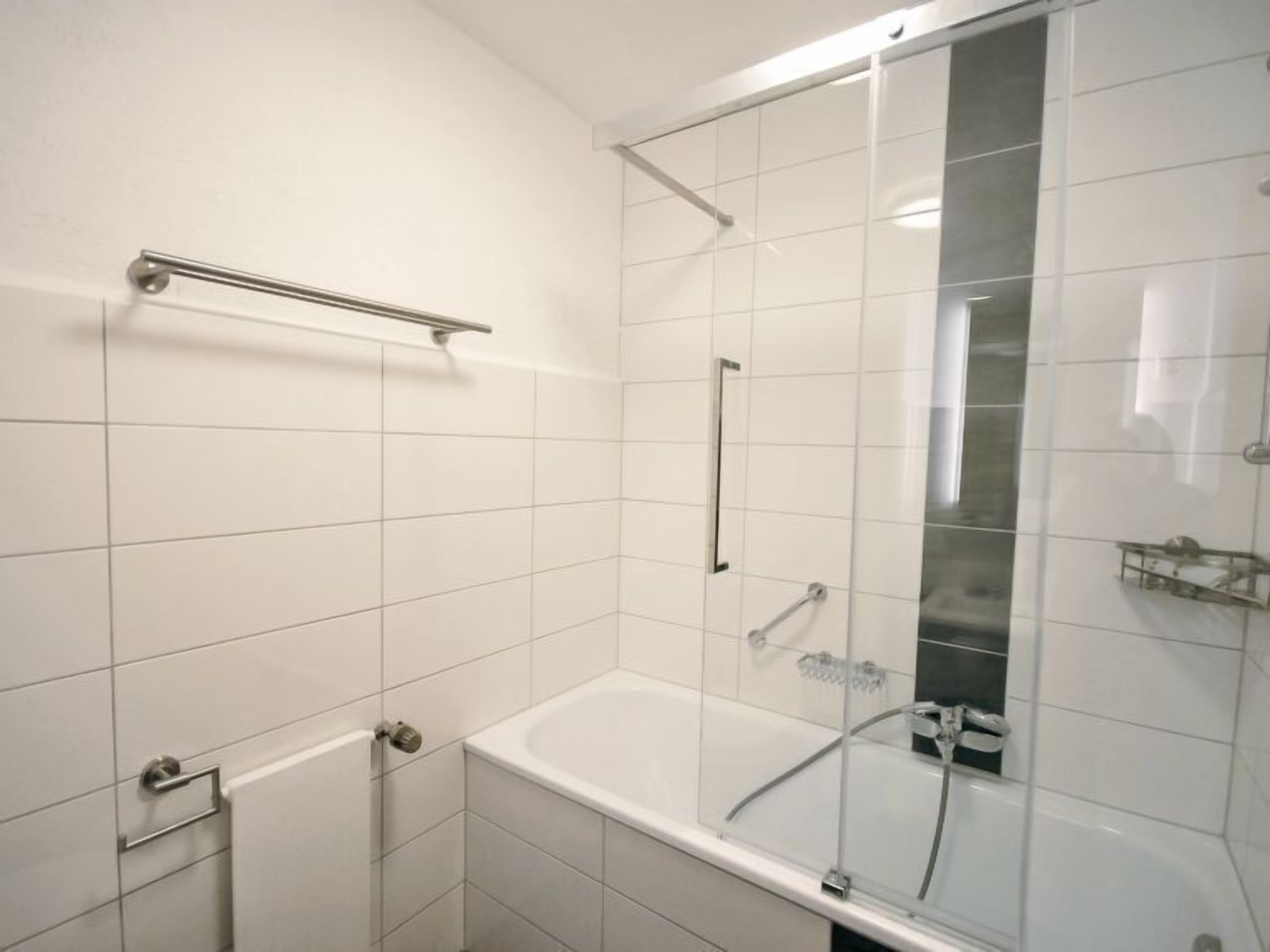 Een moderne badkamer met witte tegels, een glazen douchecabine, een badkuip en een handdoekhouder.
