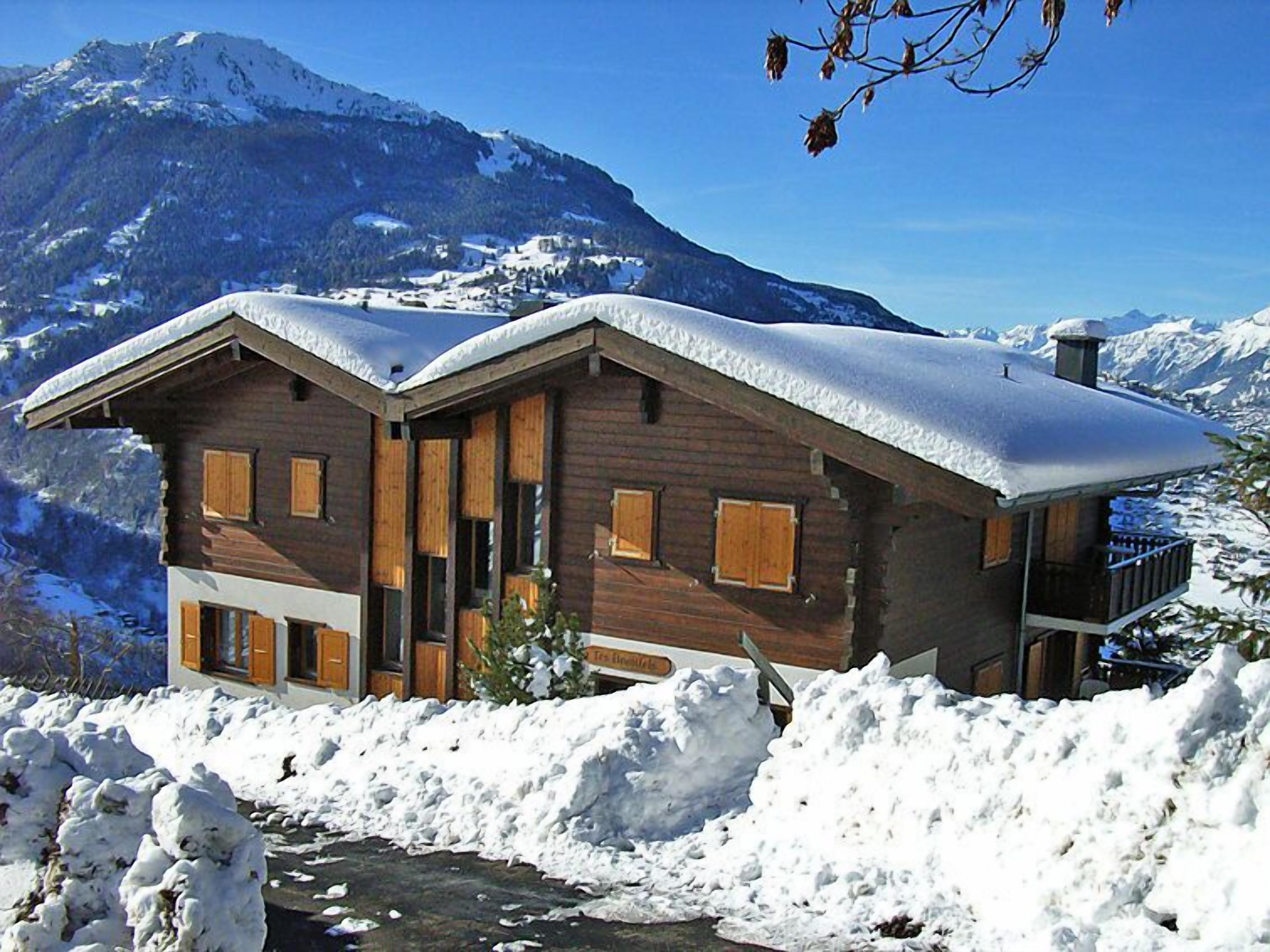 Una cabaña de madera con techo nevado, con montañas al fondo.