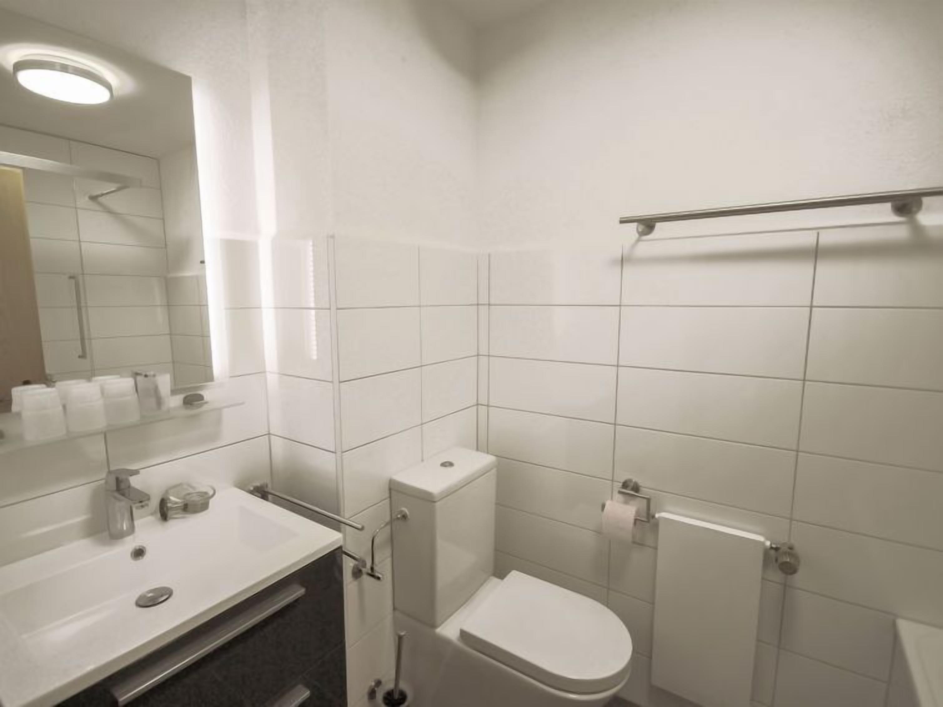 A modern bathroom with white tiles, a sink with a mirror, a toilet, and a shower area.
