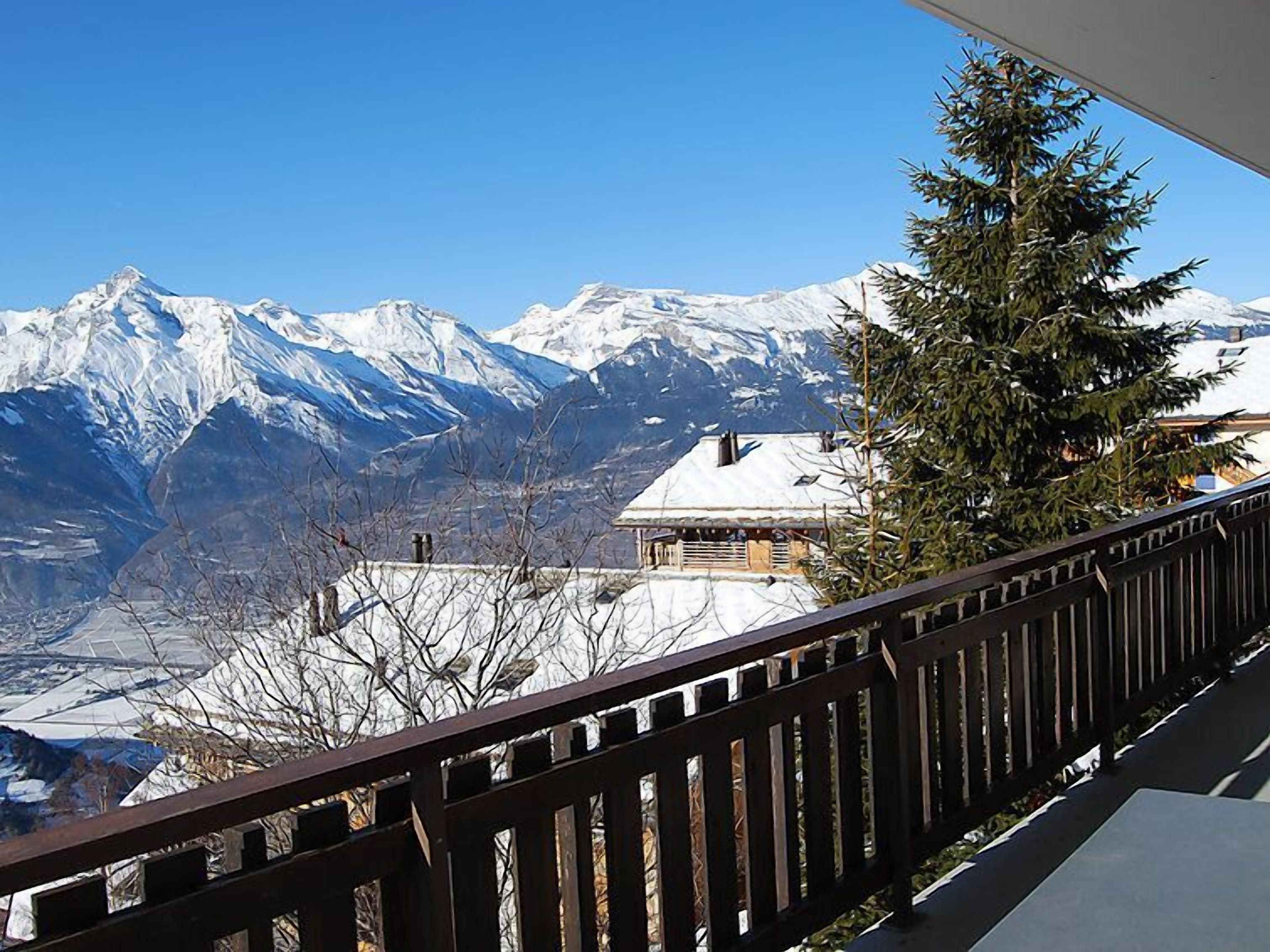 Paesaggio montano con cime innevate, un edificio in legno con tetto imbiancato dalla neve e una ringhiera di balcone in legno.