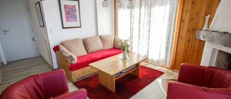 Un salon avec un canapé, une table basse et deux fauteuils rouges. Il y a une porte et une fenêtre avec des rideaux.