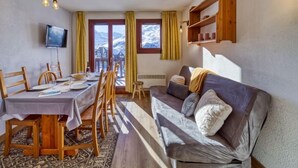 Un cuarto acogedor con una mesa de comedor lista para cuatro personas, un sofá con cojines, un televisor montado en la pared y vistas a montañas nevadas a través de las ventanas.