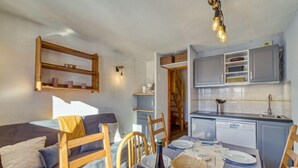Habitación pequeña con cama, una mesa de comedor para dos, zona de cocina y un estante de madera.