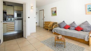 Un espace de vie compact comprenant un coin cuisine, un canapé avec des coussins, une petite table en bois et un tapis à motifs.