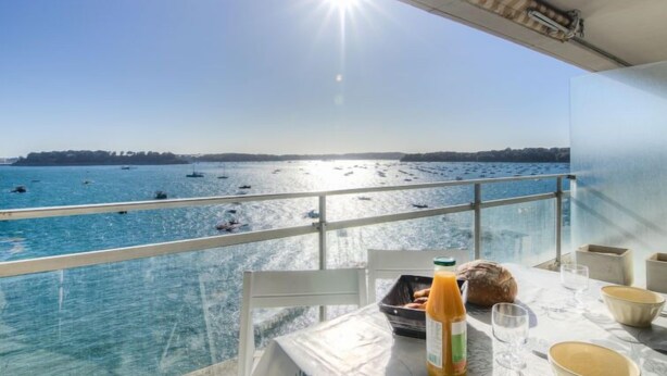 Un balcon avec une table dressée pour le petit-déjeuner, offrant une vue ensoleillée sur la côte, avec des bateaux et un ciel dégagé.