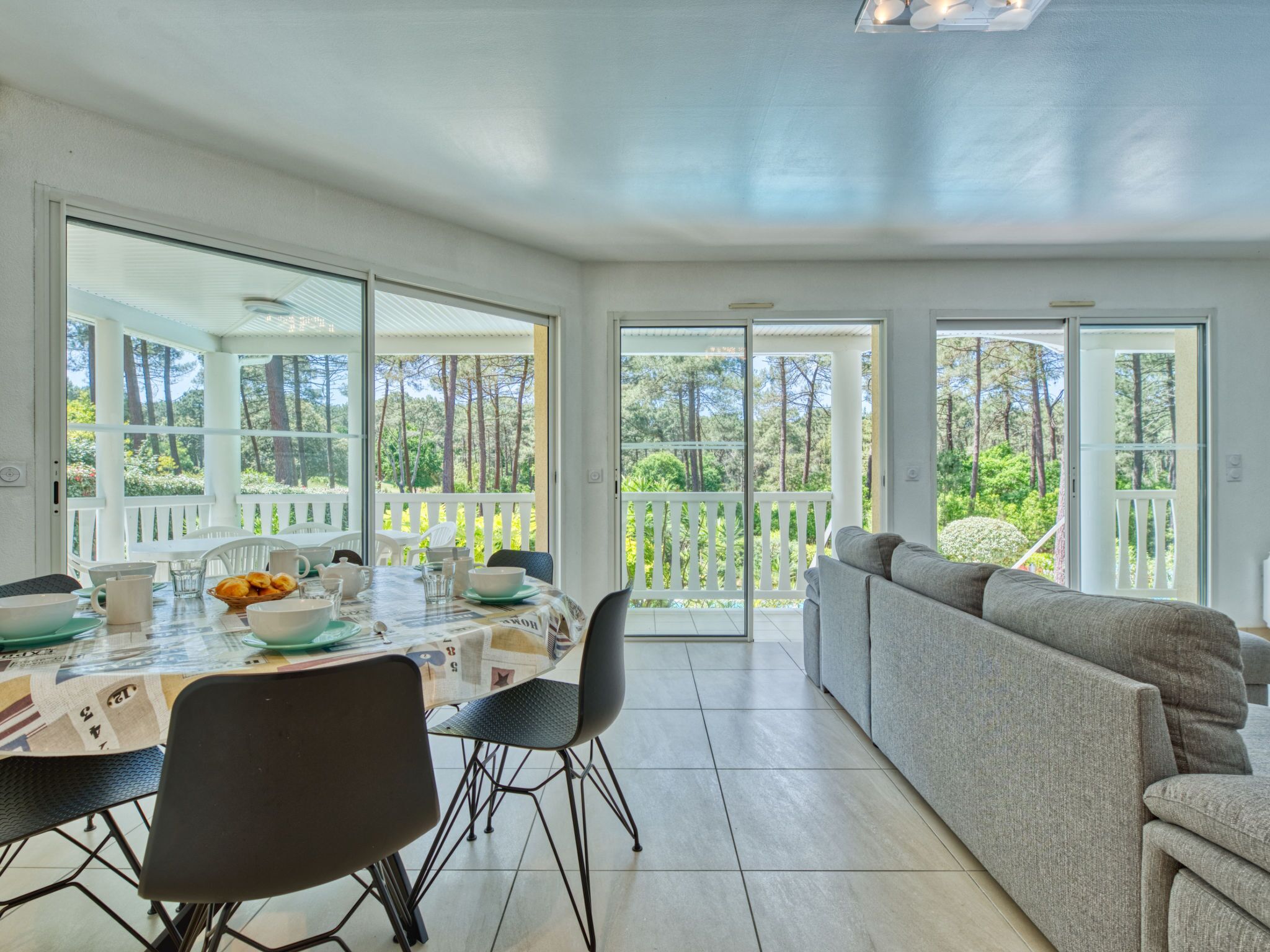 A bright, open-plan living area with a dining table set for four, a sofa, and a view of a forested area through large sliding glass doors.