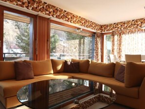 A modern living room with a large L-shaped sofa, a glass coffee table, and large windows offering a view of the outdoors.