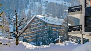 Un edifici de diversos pisos amb balcons, envoltat d'arbres i muntanyes coberts de neu.