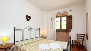 A bedroom with a bed, two bedside tables with lamps, a wooden chair, a window with curtains, and a heart-shaped decoration on the wall. - Cerbiatto n°5A by Interhome (Gaiole in Chianti)