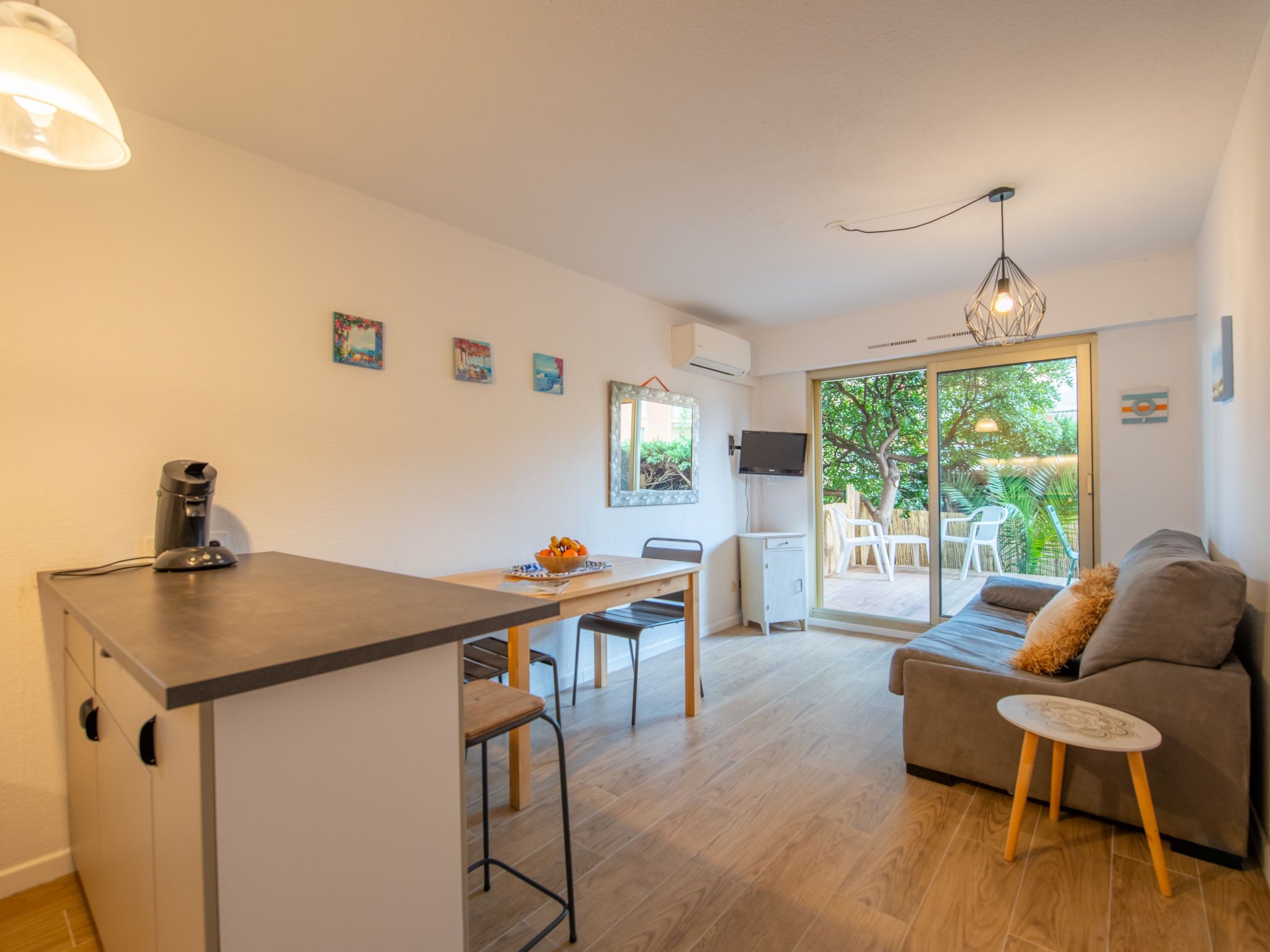 A modern kitchen with a dining area, a sofa, and a small table. There is a coffee machine on the counter and a TV mounted on the wall.