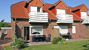 Une maison à deux étages, construite en briques, avec un toit recouvert de tuiles rouges, un balcon au deuxième étage et un petit patio aménagé avec du mobilier d’extérieur.