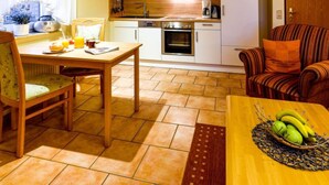 Une cuisine compacte avec des meubles blancs, un four à micro-ondes et une machine à café. On y trouve une table à manger avec des chaises, et un panier de fruits posé sur la table.