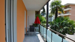 Un balcón con mesa y sillas, una sombrilla roja, y vistas a palmeras y edificios.