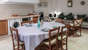 On aperçoit un coin repas avec une table ronde dressée pour six personnes, entourée de chaises en bois. Au fond, on distingue un lave-linge, et dans l’espace salon, il y a un canapé avec des coussins.