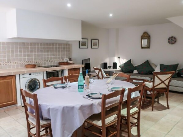 Comedor con una mesa redonda dispuesta para seis personas, rodeada de sillas de madera. Se ve una lavadora al fondo y un sofá con cojines en la sala.