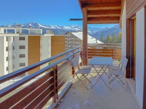 Un balcone con ringhiera in legno, un tavolo e due sedie. Il balcone si affaccia su una catena montuosa innevata.