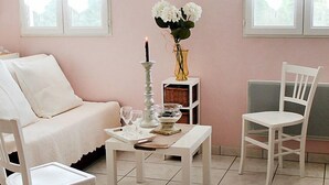 Quarto com sofá branco, mesa branca com um jogo de copos e uma cadeira branca, além de um vaso com flores.