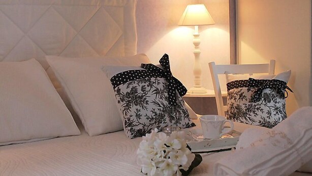 Cama bem arrumada com lençóis brancos, duas almofadas com estampas florais pretas, uma mesa pequena com xícara e pires, e um vaso com flores brancas.