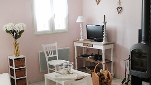 Quarto acolhedor com uma cadeira branca, uma mesinha com copos, uma televisão em um suporte branco, uma lareira e um vaso com flores.