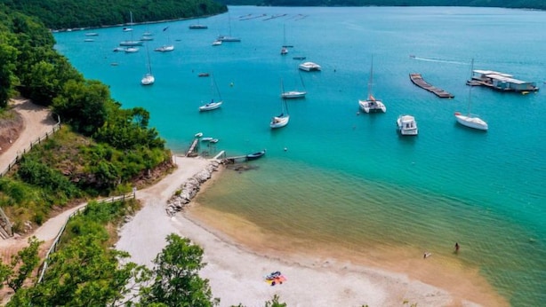 Zona costera con barcos amarrados en aguas tranquilas, rodeada de vegetación y colinas.