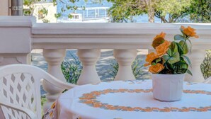 Un balcón con barandilla blanca, una mesa con mantel floral y una maceta con flores naranjas.