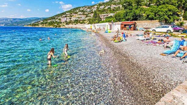 Una playa de guijarros con agua cristalina, gente nadando y una colina con edificios.