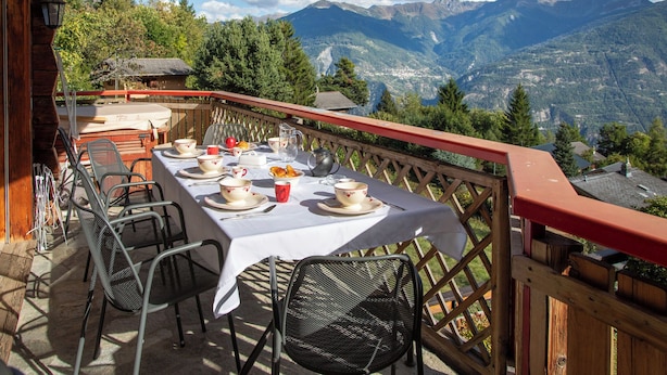 Taula preparada per esmorzar amb vista a la muntanya, cadira exterior i barana de fusta.
