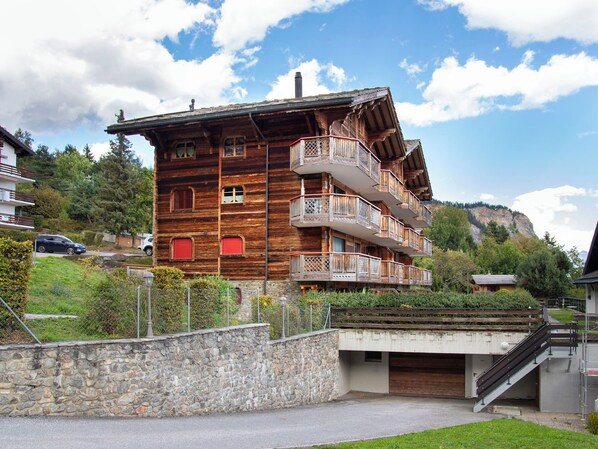 A multi-story wooden building with balconies, a stone wall, and a mountain backdrop.