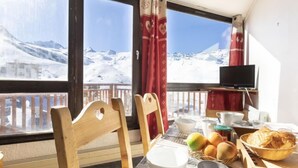 Uma área de refeições com uma mesa posta para o café da manhã, uma televisão em um suporte e vista para montanhas cobertas de neve através de grandes janelas.