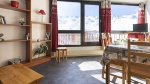 Quarto aconchegante com estante de madeira, mesa pequena e área de refeições com cadeiras. Uma janela oferece vista para montanhas nevadas.