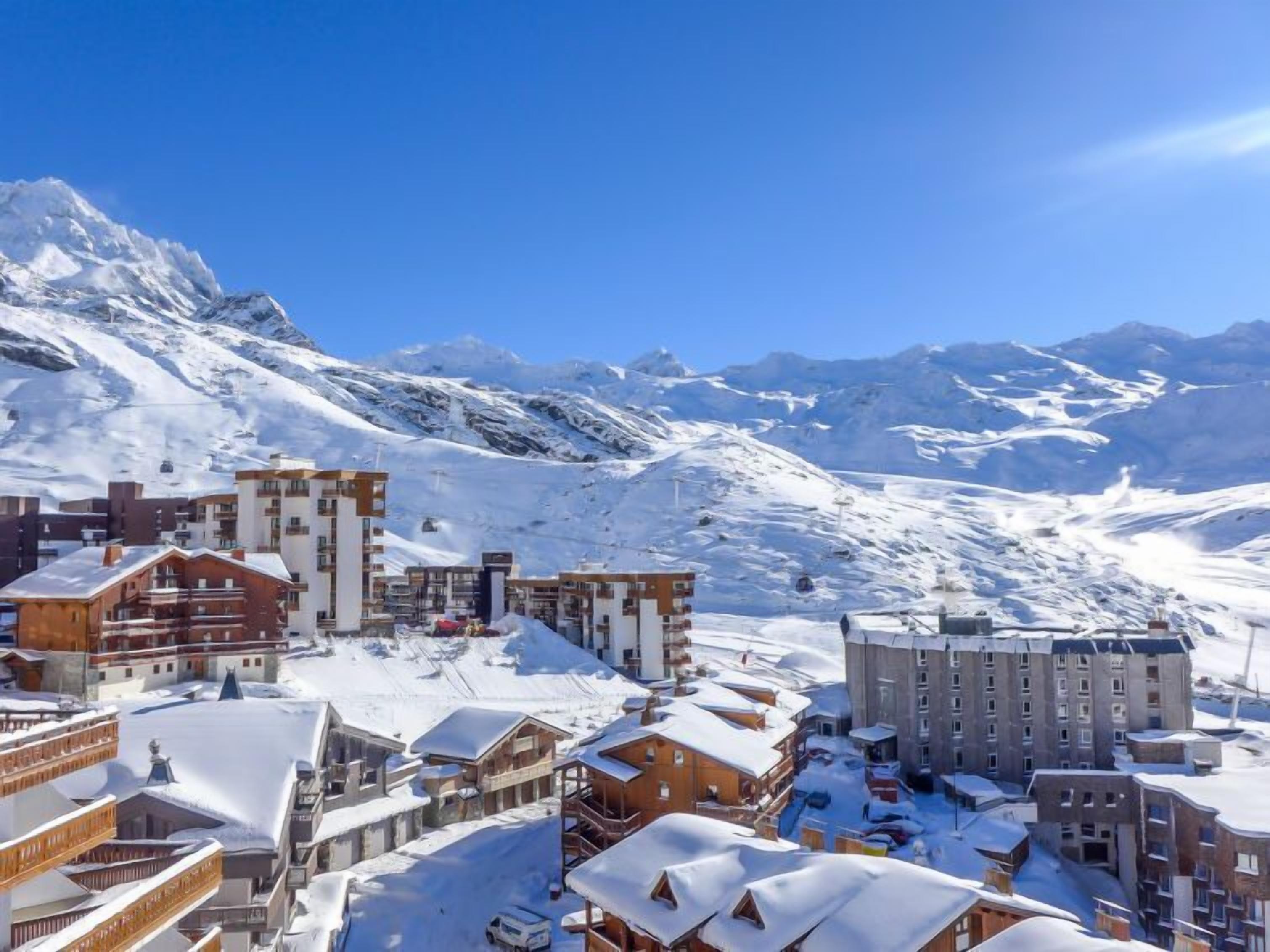 Uma cidade montanhosa coberta de neve, com edifícios modernos, incluindo um hotel grande com vários andares e varandas, com picos nevados ao fundo.