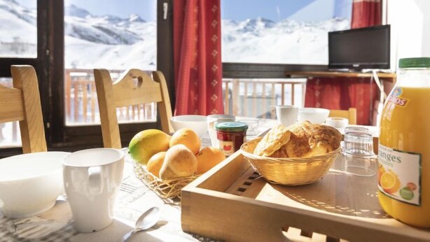 Mesa de café da manhã com pão, frutas e uma garrafa de suco em um quarto com vista para as montanhas.