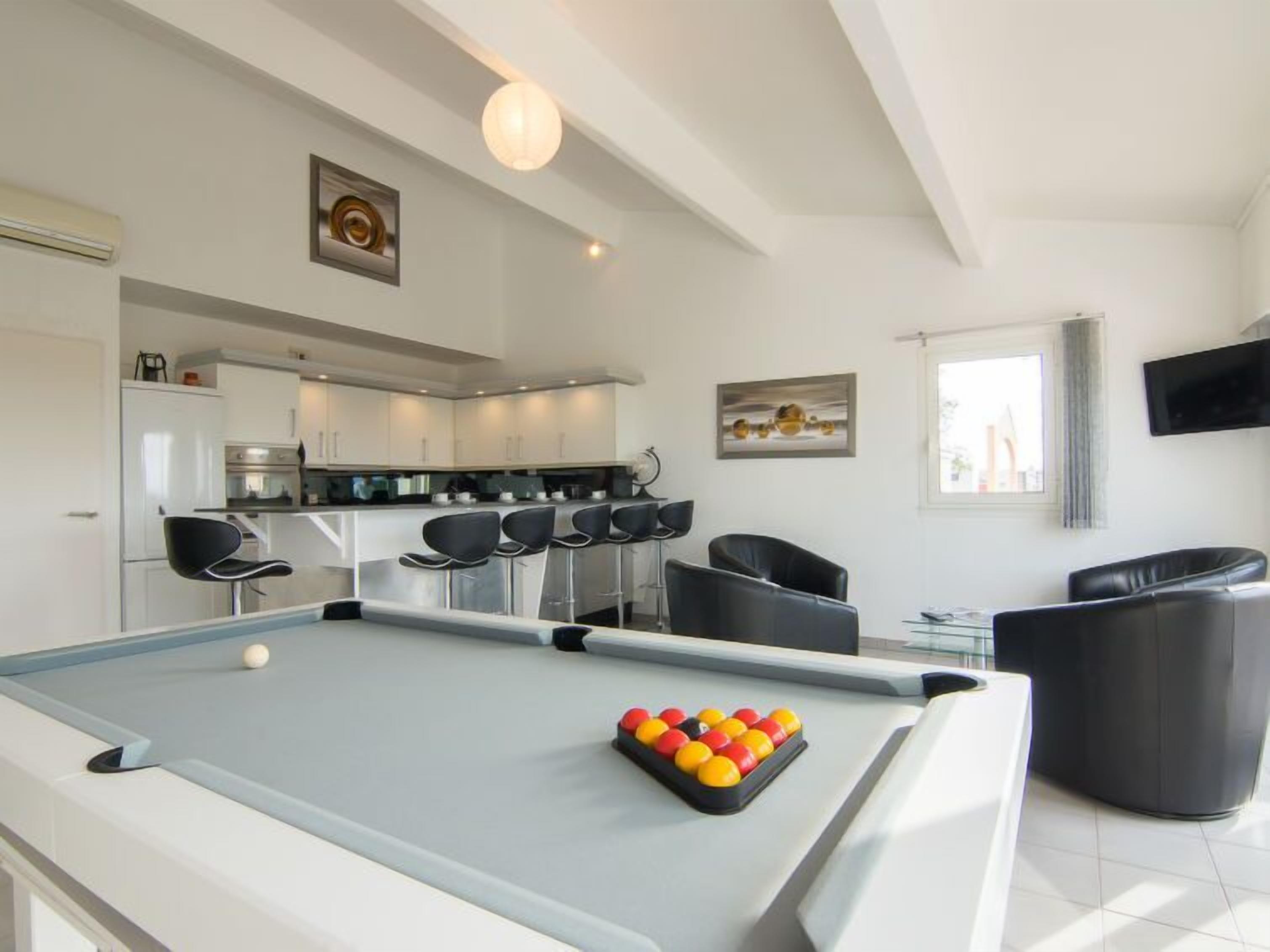 A modern living room with a pool table, black leather chairs, a kitchen area, and artwork on the walls.