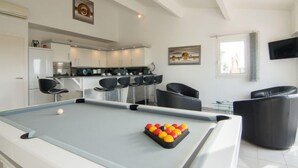 Ein modernes Wohnzimmer mit Billardtisch, schwarzen Ledersesseln, einer Küchenzeile und Kunstwerken an den Wänden.