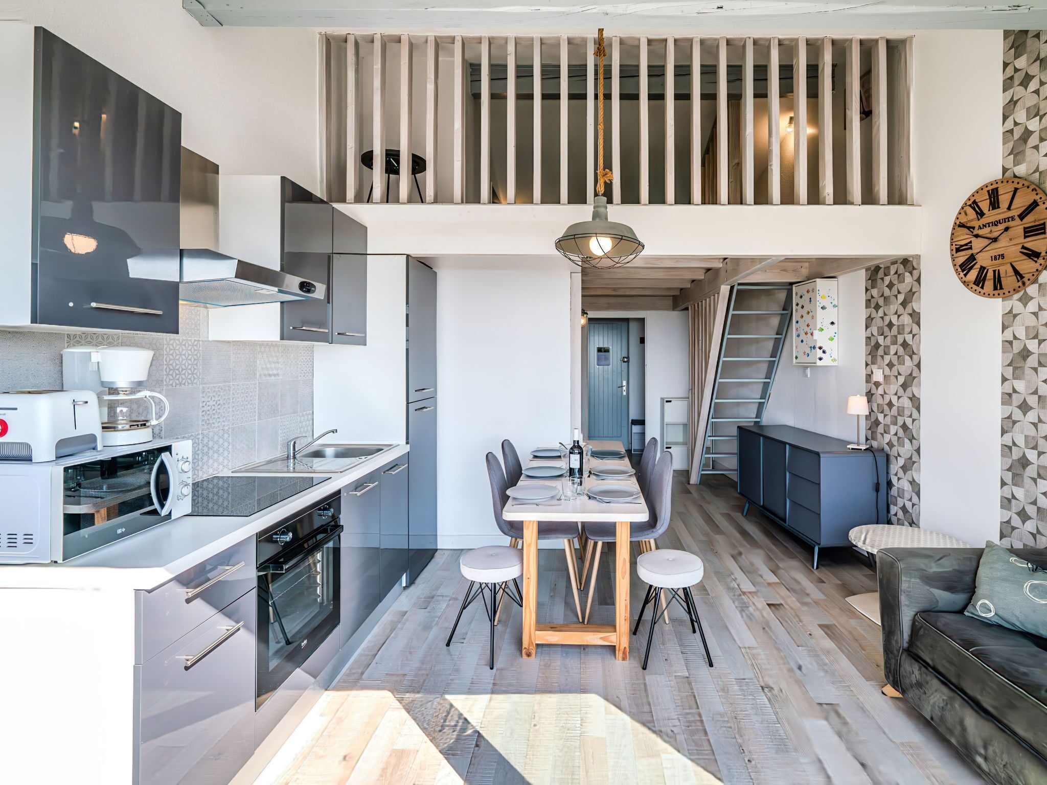 A modern kitchen with a dining area, a loft bedroom, and a living room with a sofa and a clock.