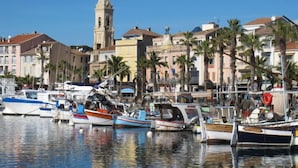 A marina with various boats docked, a prominent church tower, and buildings with balconies and palm trees.