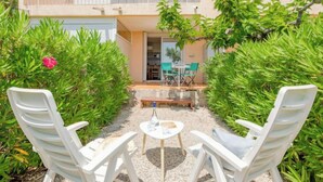 Eine Terrasse mit weißen Plastikstühlen und einem kleinen runden Tisch, umgeben von grünen Sträuchern und einer rosa Blume.