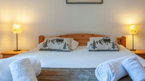 Een net opgemaakt bed met twee kussens met een bergmotief, bezaaid met twee nachtlampjes. Daarboven hangt een ingelijst berglandschap.