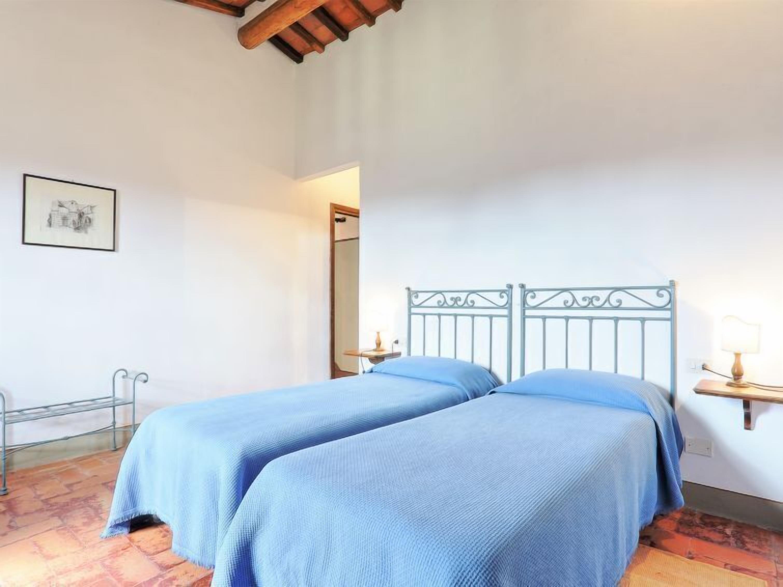 A room with two beds, a metal-framed headboard, a small bedside table with a lamp, and a framed picture on the wall.