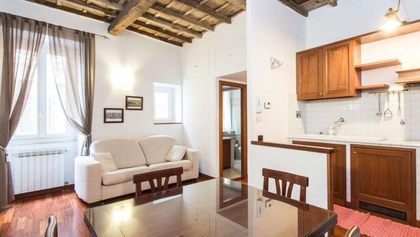 A cozy living room with a sofa, a glass dining table, wooden beams on the ceiling, and a kitchen with wooden cabinets and a white countertop. - Domus Fori by Interhome (Rome: Historical City Center)