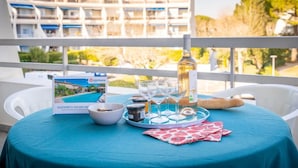 Una mesa dispuesta con una botella, copas, un bol y una bandeja con condimentos, con vista a un edificio con balcones y vegetación.