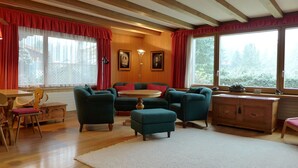 A cozy living room with wooden beams, a sofa set, a dining table, and a view of greenery outside. - Chalet Marietta by Interhome (Kandersteg)