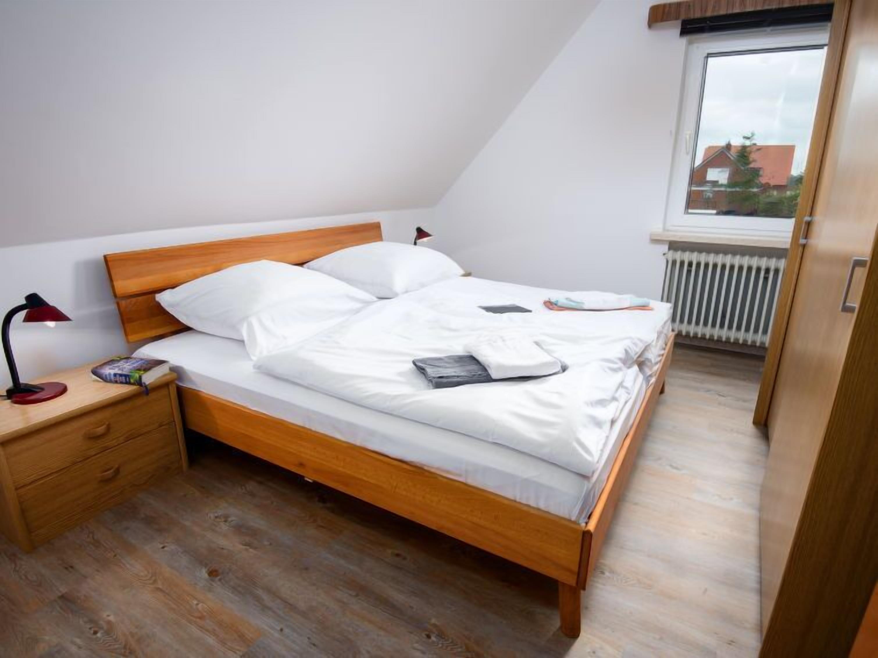 Ein Schlafzimmer mit einem hölzernen Bett, weißer Bettwäsche, einem Nachttisch mit Lampe und einem Fenster mit Blick auf Gebäude.