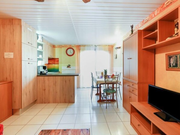 Een kleine keuken met houten kastjes, een eethoek met een tafel en stoelen, en een televisie op een houten dressoir.