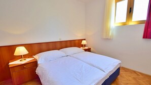 Un dormitorio con cabecera de madera, dos mesitas de noche con lámparas, una cama grande con ropa blanca y una ventana con cortinas.