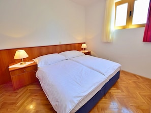 Ein Schlafzimmer mit Holz-Kopfteil, zwei Nachttischen mit Lampen, einem großen Bett mit weißer Bettwäsche und einem Fenster mit Vorhängen.