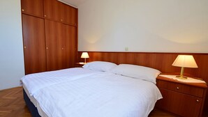 Een opgemaakt bed met witte lakens, twee nachtkastjes met lampen en een houten garderobe.