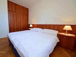 Una cama bien tendida con sábanas blancas, dos mesitas de noche con lámparas y un armario de madera.