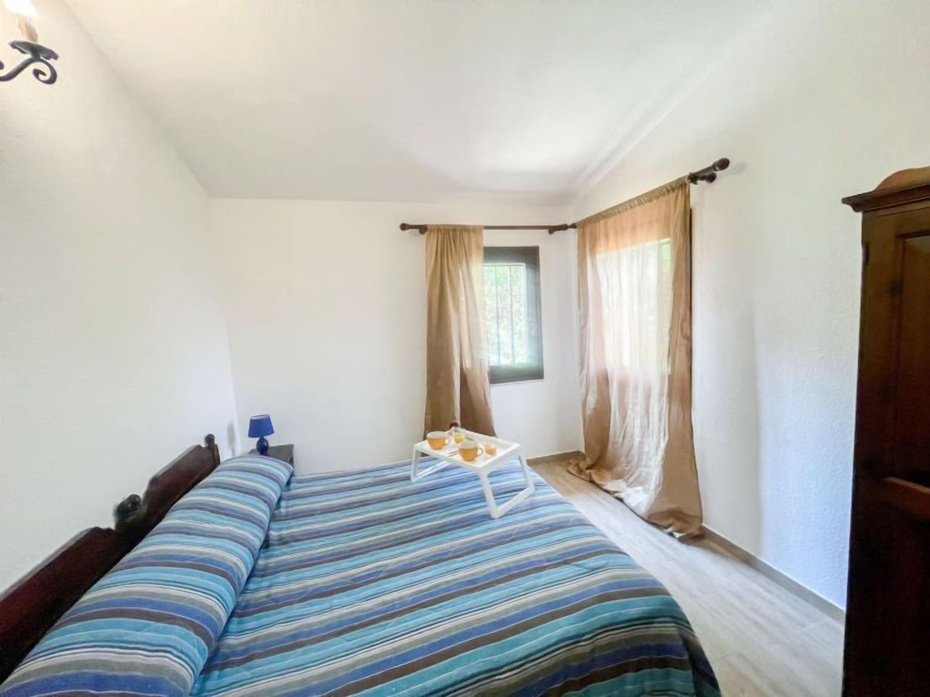 A single-bed room with a striped bedspread, a bedside table with a lamp, and a window with curtains.