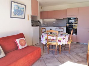 A compact living space with a red sofa, a dining table with chairs, and a kitchen area with pink cabinets and appliances. - Les Romarines by Interhome (Gruissan)